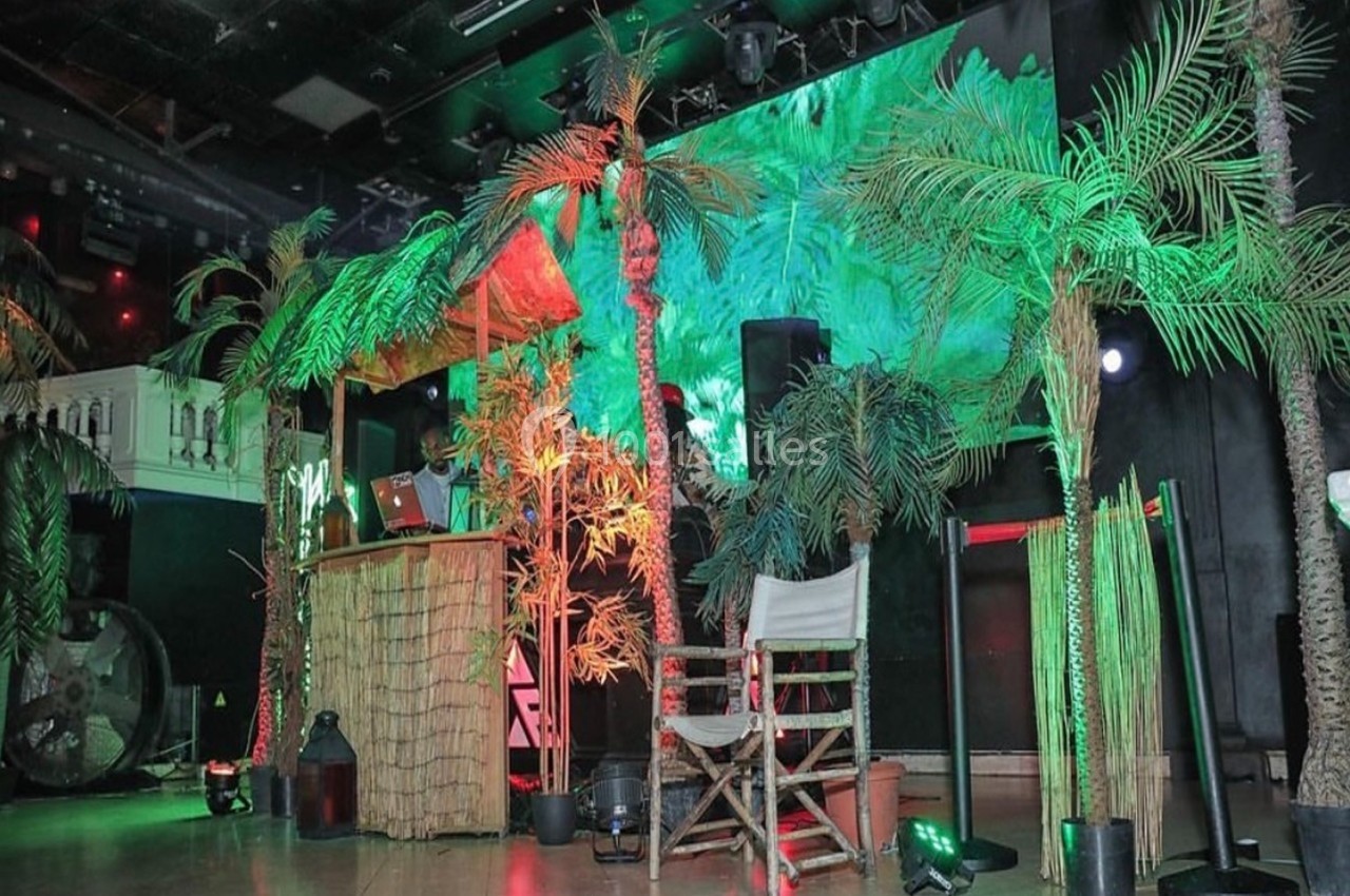 Décor tropical avec palmiers, cabane en bois et éclairages colorés dans une salle sombre.