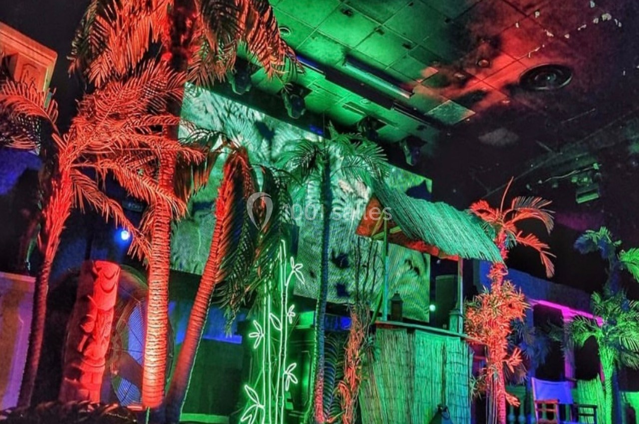 Décor tropical intérieur avec palmiers artificiels, éclairages colorés et cabane en bois sous un plafond sombre.