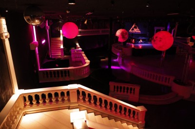 Intérieur d'une discothèque avec éclairage violet, escalier central, balustrades métalliques et sol carrelé coloré.