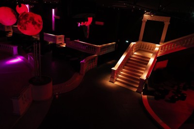 Intérieur d'une discothèque avec éclairage violet, escalier central, balustrades métalliques et sol carrelé coloré.