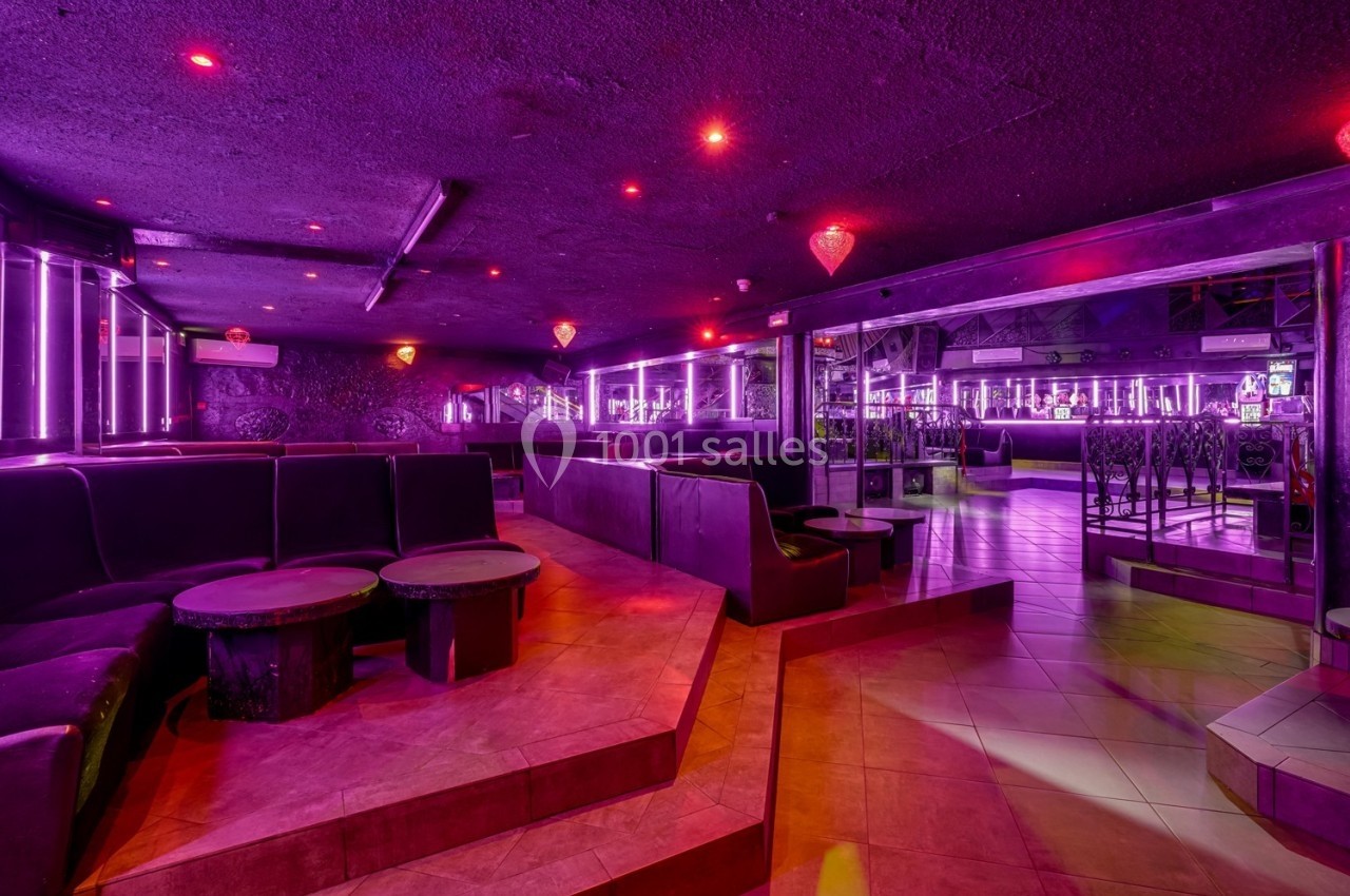 Intérieur d'un club avec éclairage violet, banquettes, tables basses et sol carrelé.