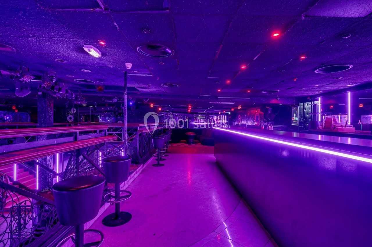 Salle de discothèque vide avec éclairage violet, comptoir lumineux et tabourets alignés le long d'une rambarde.