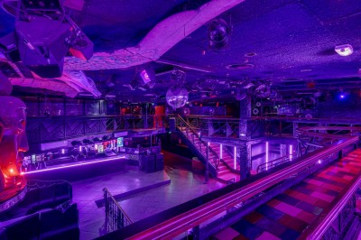 Intérieur d'une discothèque avec éclairage violet, escalier central, balustrades métalliques et sol carrelé coloré.