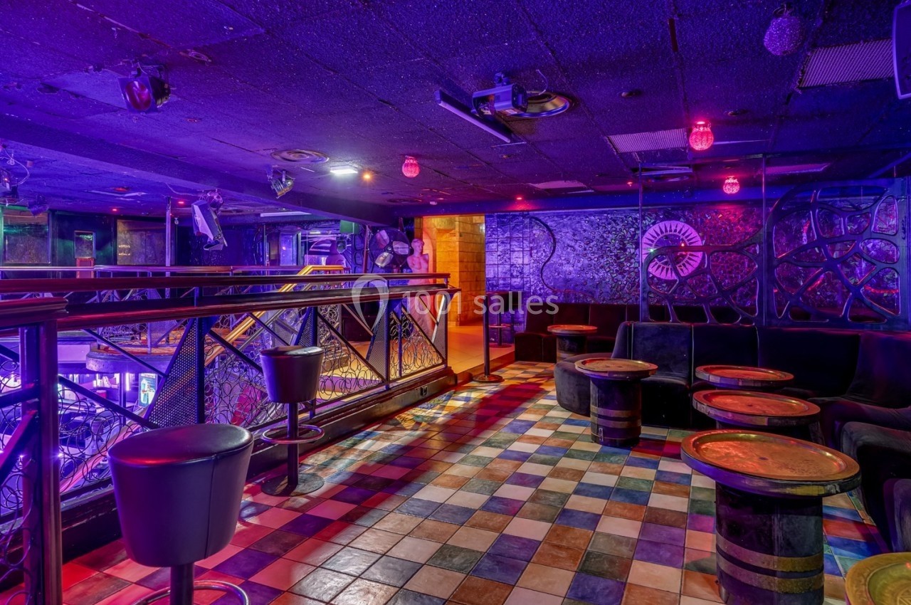 Salle de bar colorée avec tables rondes, sièges en cuir noir et éclairage violet dans une ambiance nocturne.