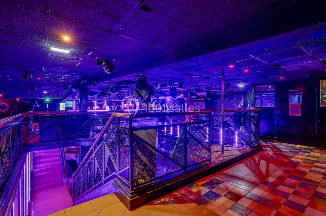 Intérieur d'une discothèque avec éclairage violet, escalier central, balustrades métalliques et sol carrelé coloré.