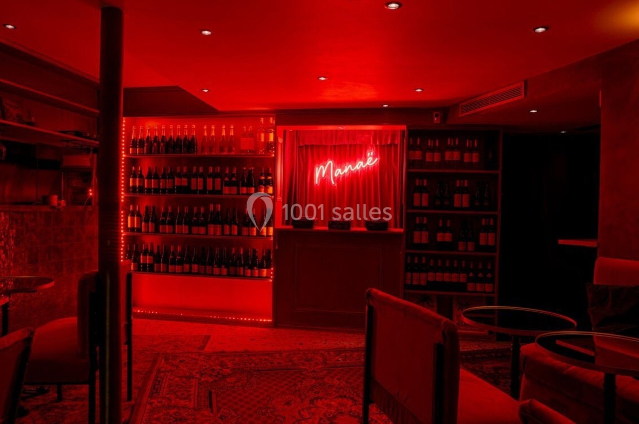 Bar à l'éclairage rouge tamisé avec étagères de bouteilles, tables basses et enseigne lumineuse ’Maria’.