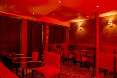 Salle intimiste éclairée par une lumière rouge, avec banquettes, petites tables rondes et rideaux sombres.