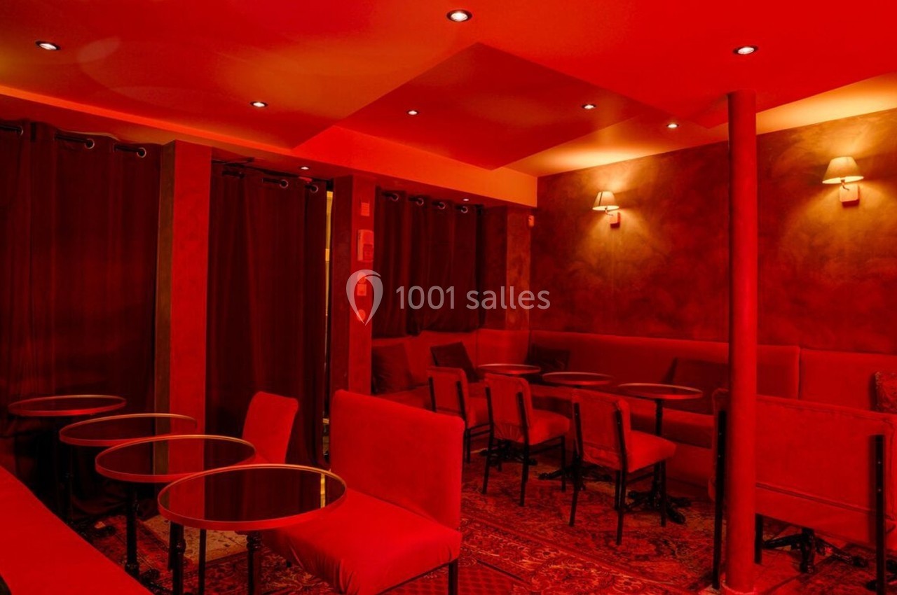 Salle intimiste éclairée de lumière rouge, avec des tables rondes, des chaises et des rideaux sombres.