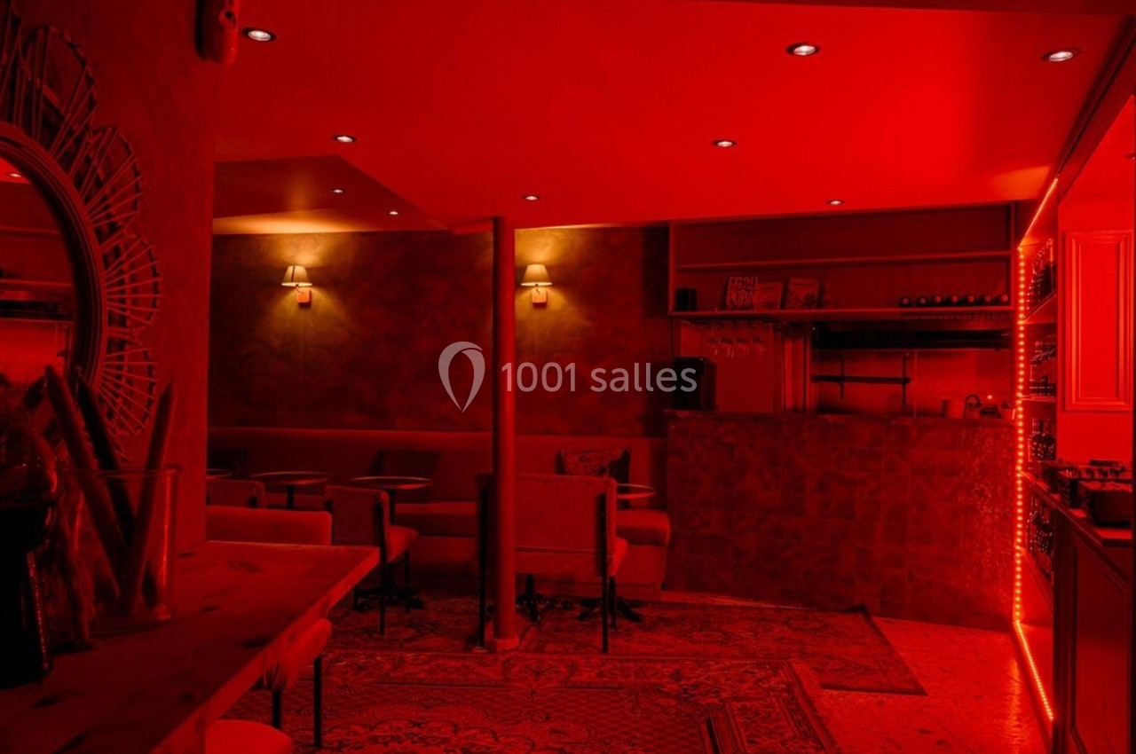 Salle de bar à l'éclairage rouge tamisé, avec des tables, des chaises et des étagères de bouteilles.