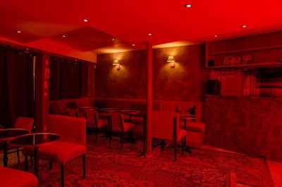 Salle intimiste éclairée par une lumière rouge, avec banquettes, petites tables rondes et rideaux sombres.