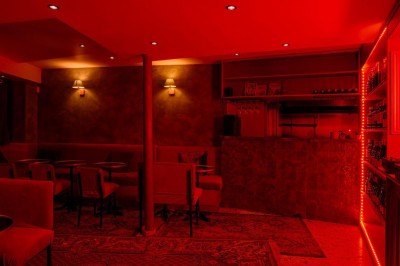 Salle intimiste éclairée par une lumière rouge, avec banquettes, petites tables rondes et rideaux sombres.