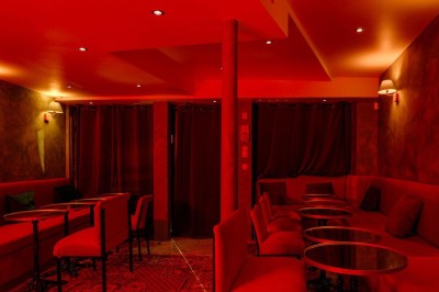 Salle intimiste éclairée par une lumière rouge, avec banquettes, petites tables rondes et rideaux sombres.
