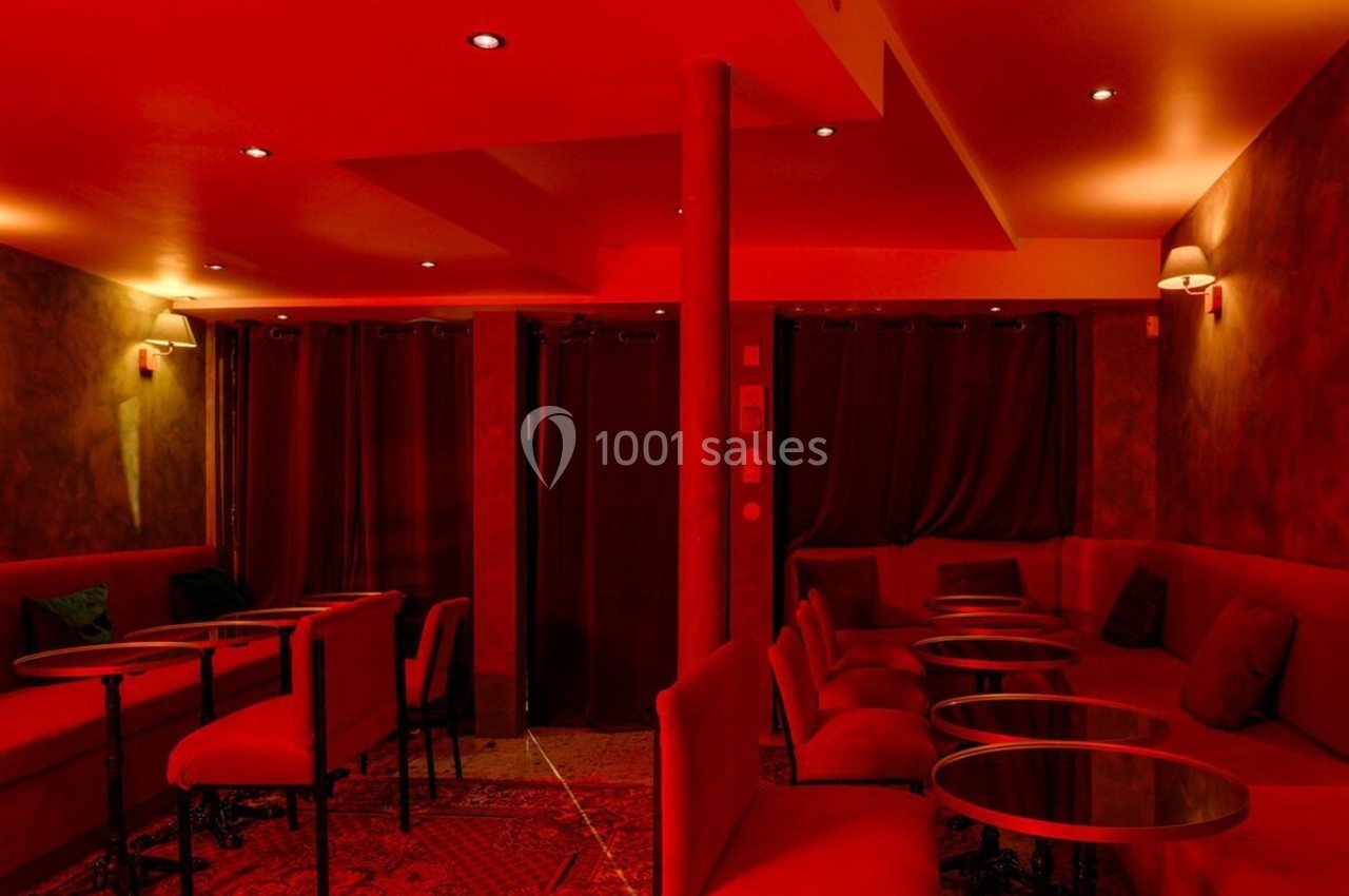 Salle intimiste éclairée de lumière rouge, avec canapés, petites tables rondes et rideaux sombres en arrière-plan.