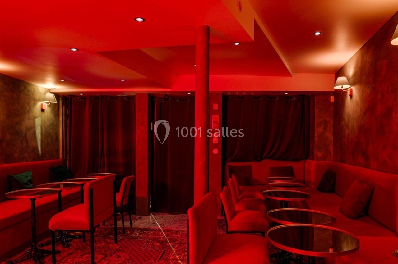 Salle intimiste éclairée par une lumière rouge, avec banquettes, petites tables rondes et rideaux sombres.
