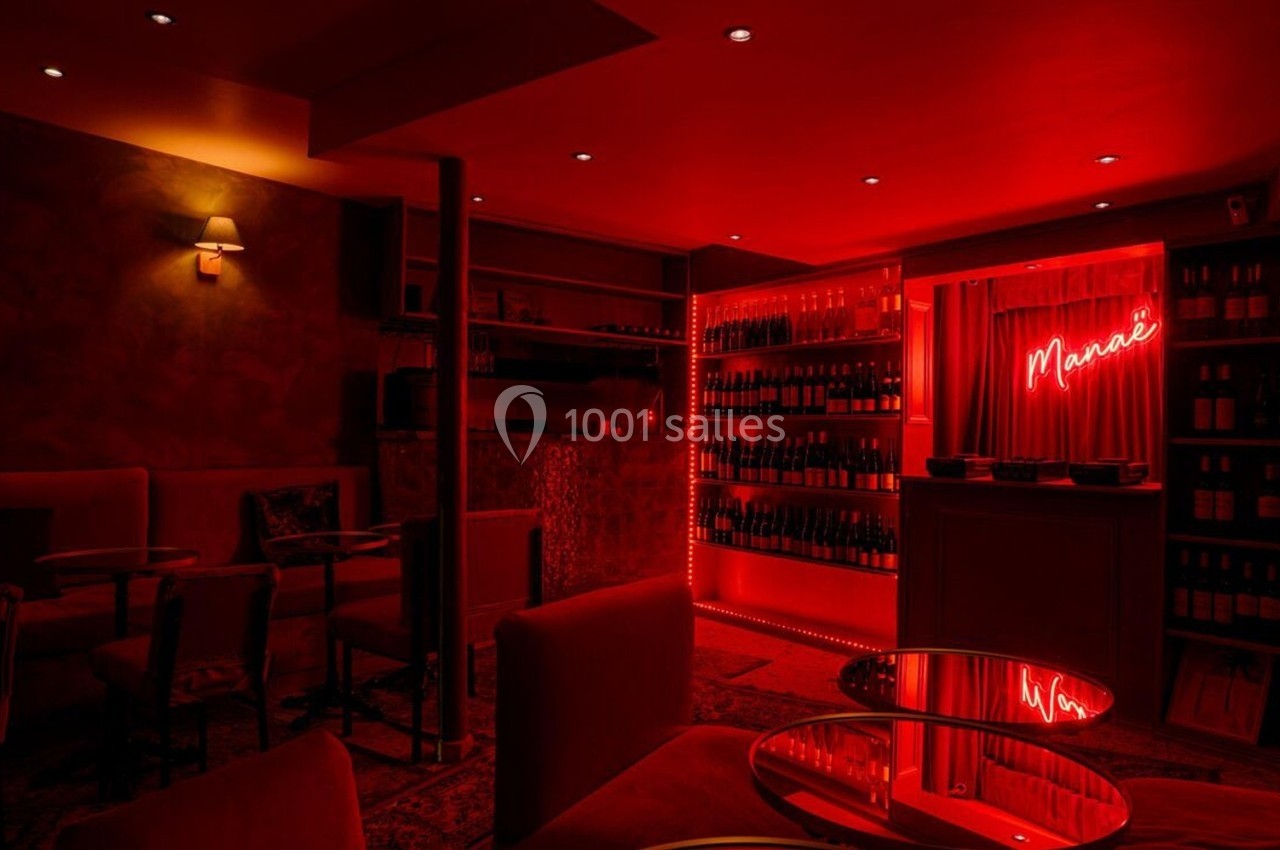 Salle de bar intimiste éclairée par une lumière rouge, avec des étagères de bouteilles et un néon ’Merci’.