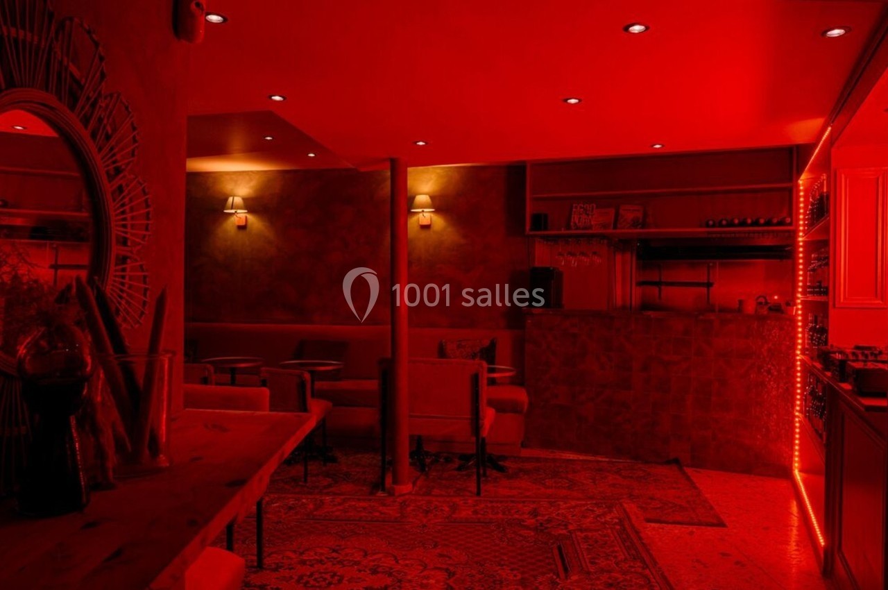 Salle intimiste éclairée par une lumière rouge, avec des fauteuils, un bar et des étagères de bouteilles.