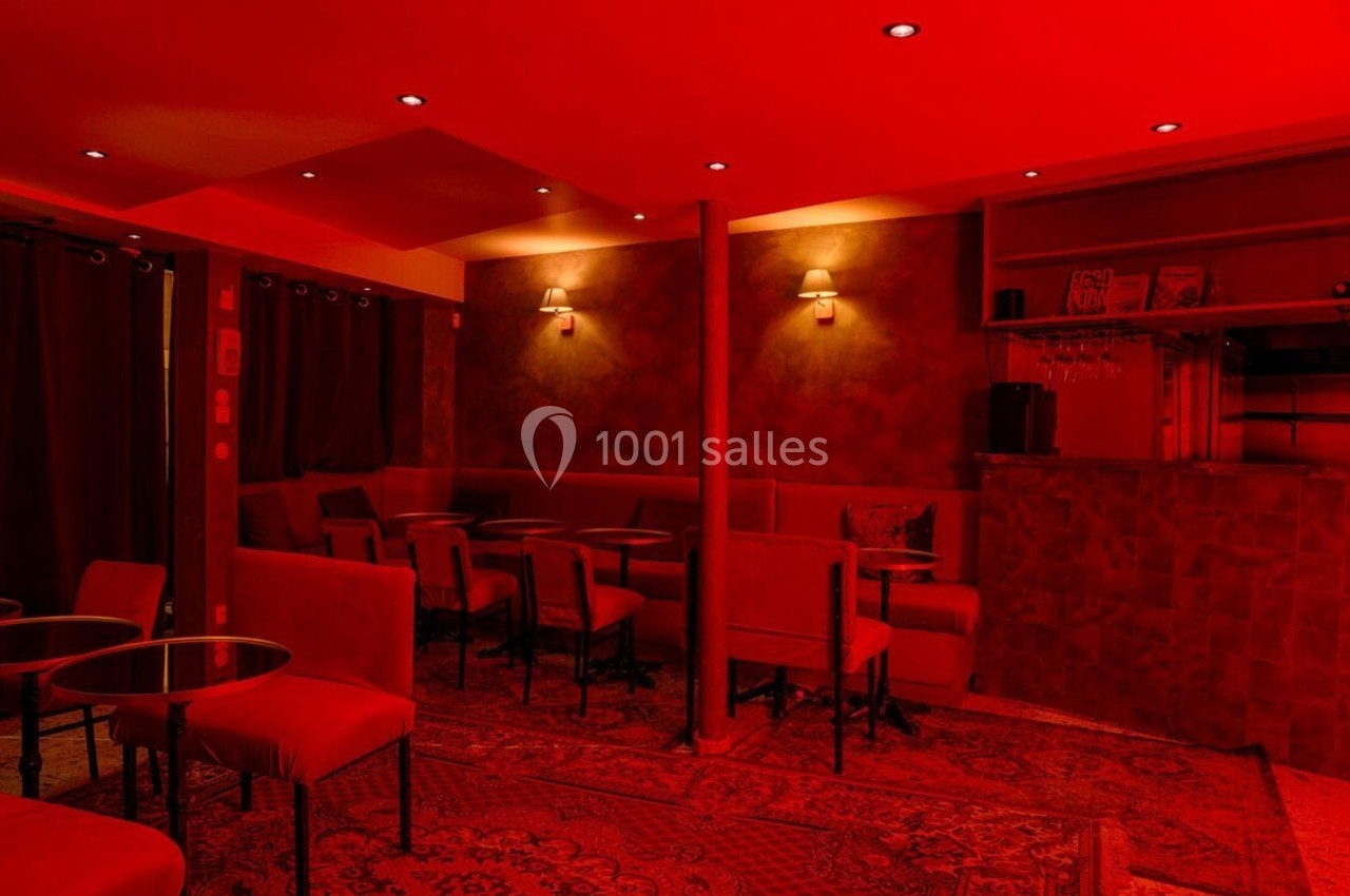 Salle de bar intimiste éclairée de lumières rouges, avec tables, chaises et comptoir en arrière-plan.