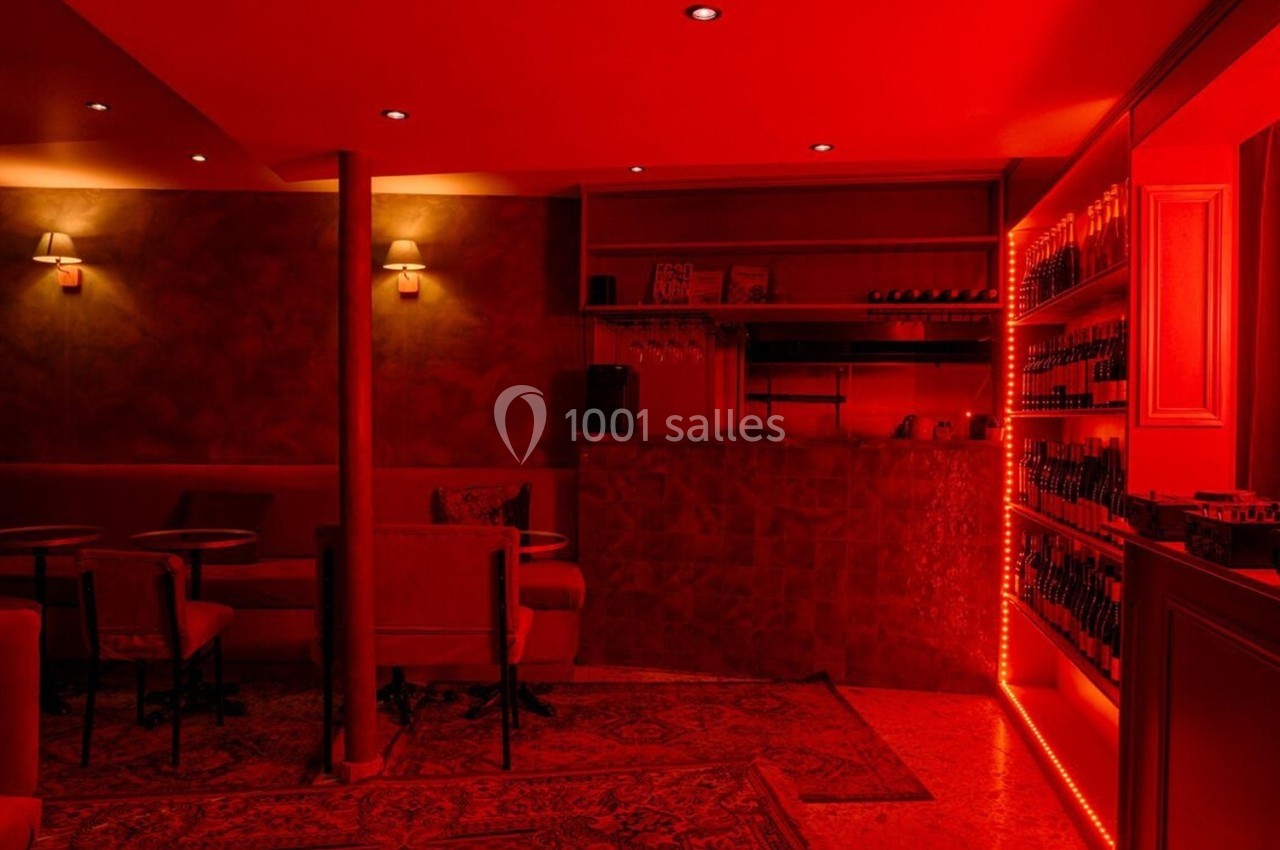 Salle de bar à l'éclairage rouge tamisé, avec tables, chaises et étagères remplies de bouteilles.