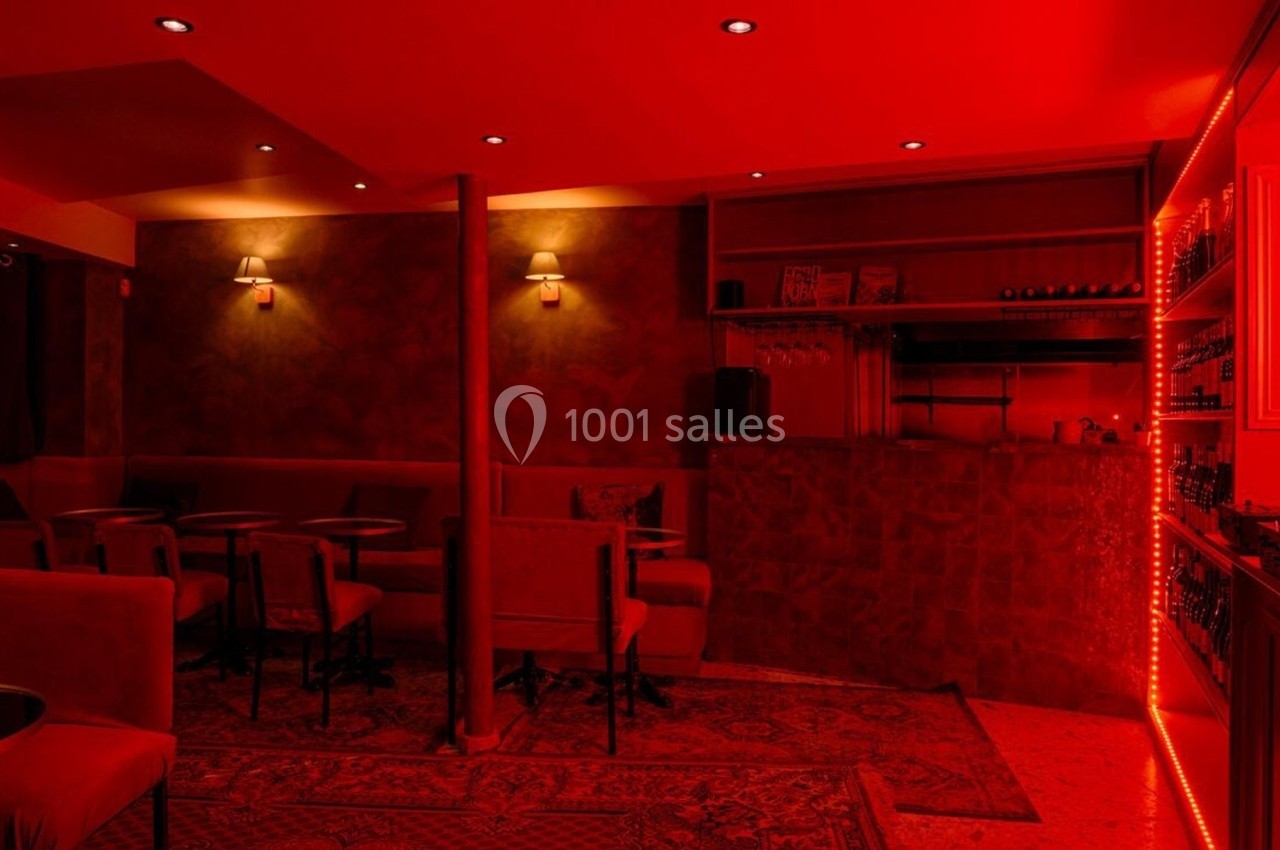 Salle de bar à l'éclairage rouge, avec tables, chaises, comptoir et étagères garnies de bouteilles.