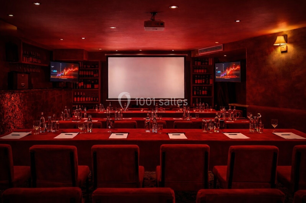 Salle de conférence sombre avec écran principal, deux écrans latéraux, tables rouges, bouteilles d'eau et documents.