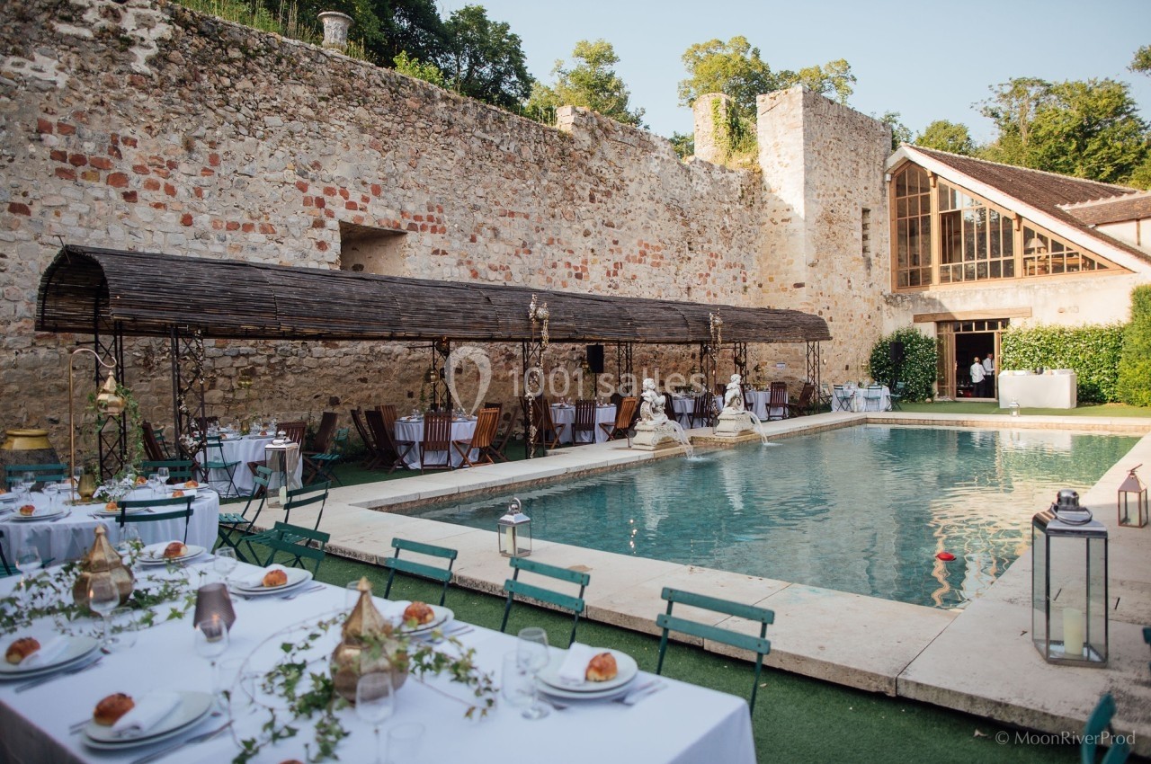Terrasse avec tables dressées autour d'une piscine, devant un mur en pierre et une bâtisse avec grandes fenêtres.