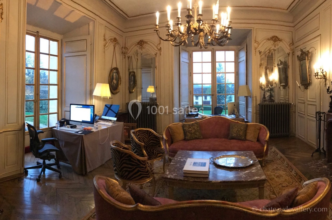 Salon élégant avec moulures, lustre, mobilier classique, bureau avec ordinateur et vue sur un jardin par des fenêtres.