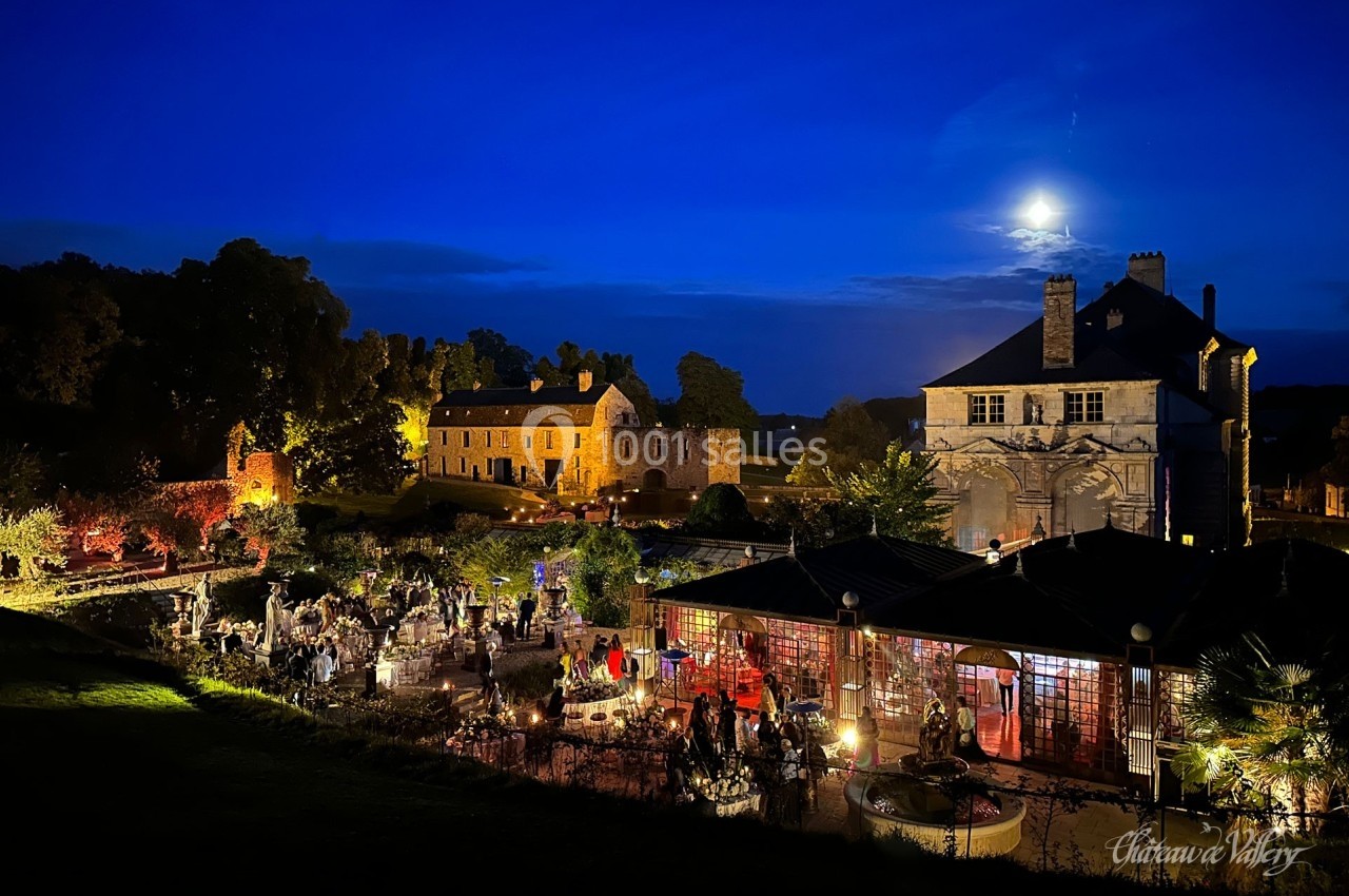 Vue nocturne d'un château éclairé avec des invités rassemblés dans un jardin illuminé sous une pleine lune.