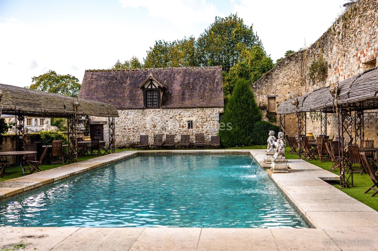Piscine extérieure entourée de bâtiments en pierre, de chaises en bois et de végétation.