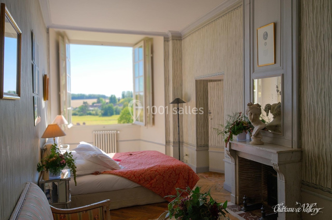 Chambre lumineuse avec lit double, cheminée, décor élégant et vue sur un paysage verdoyant par une grande fenêtre ouverte.