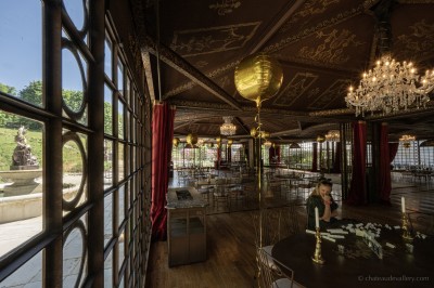 Salle lumineuse avec grandes baies vitrées, tables rondes en métal et lustres suspendus sous un plafond décoré.