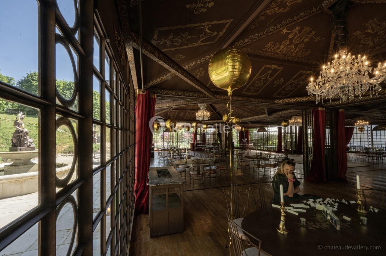 Salle élégante avec grandes baies vitrées, lustres en cristal, décor doré et vue sur une statue dans un jardin.