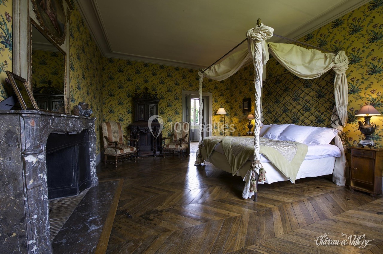 Chambre avec lit à baldaquin, parquet en bois, cheminée en marbre, murs tapissés de motifs floraux et mobilier ancien.