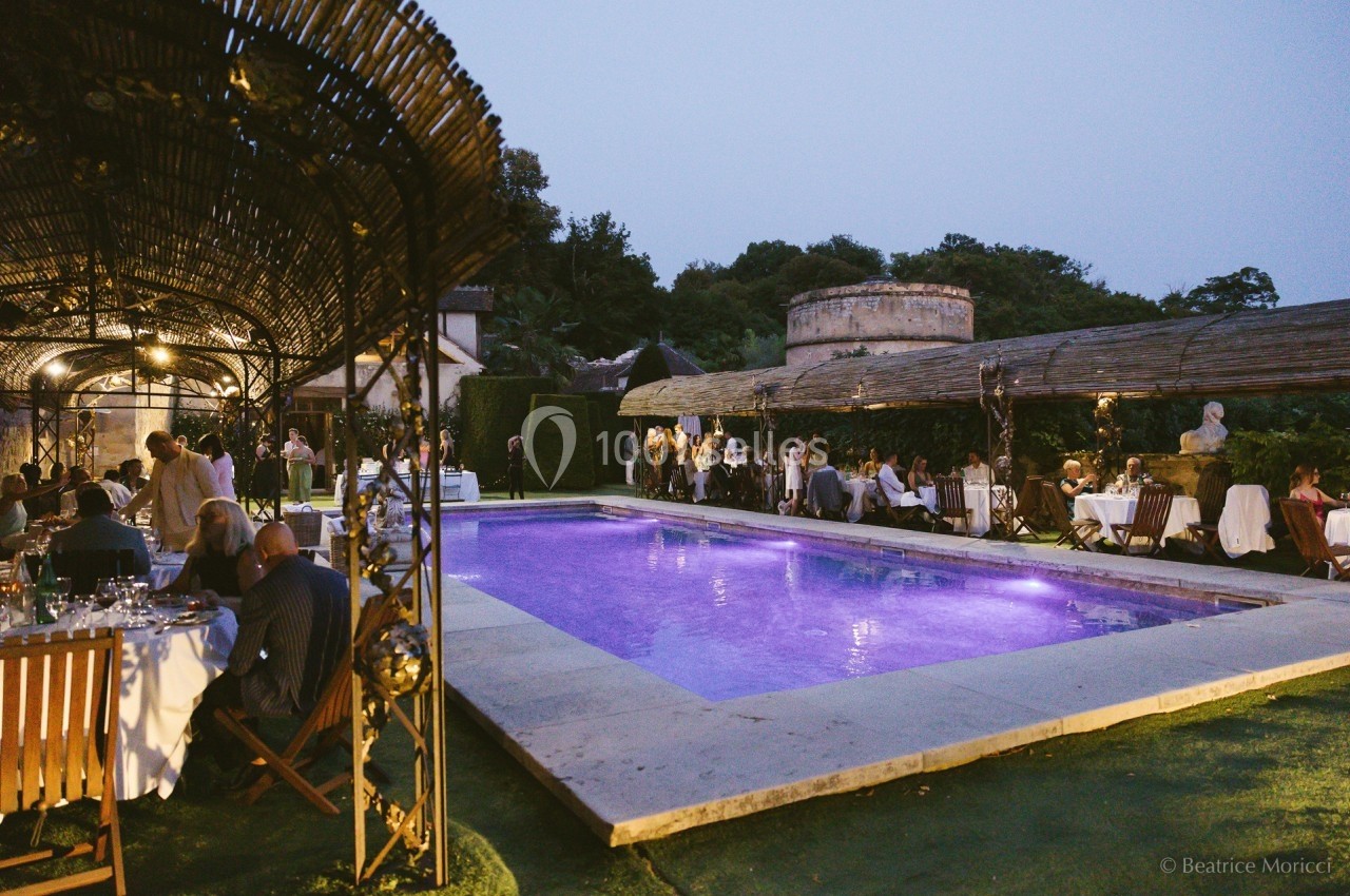 Dîner en plein air autour d'une piscine éclairée, avec des invités assis sous des pergolas au crépuscule.