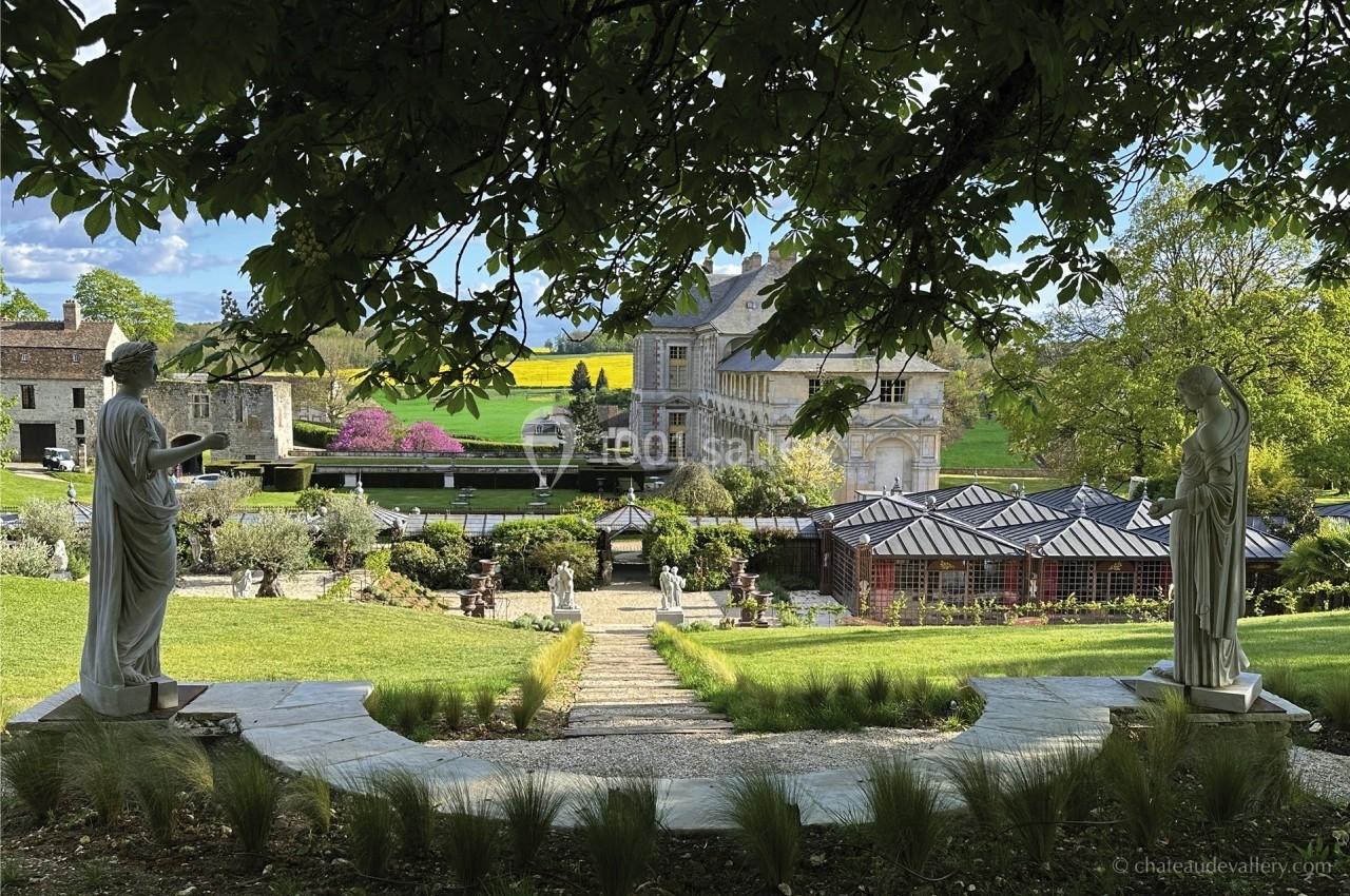 Vue d'un jardin paysager avec statues, allée centrale, serres et un bâtiment historique en arrière-plan.
