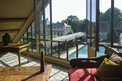 Salle lumineuse avec grandes baies vitrées, tables rondes en métal et lustres suspendus sous un plafond décoré.