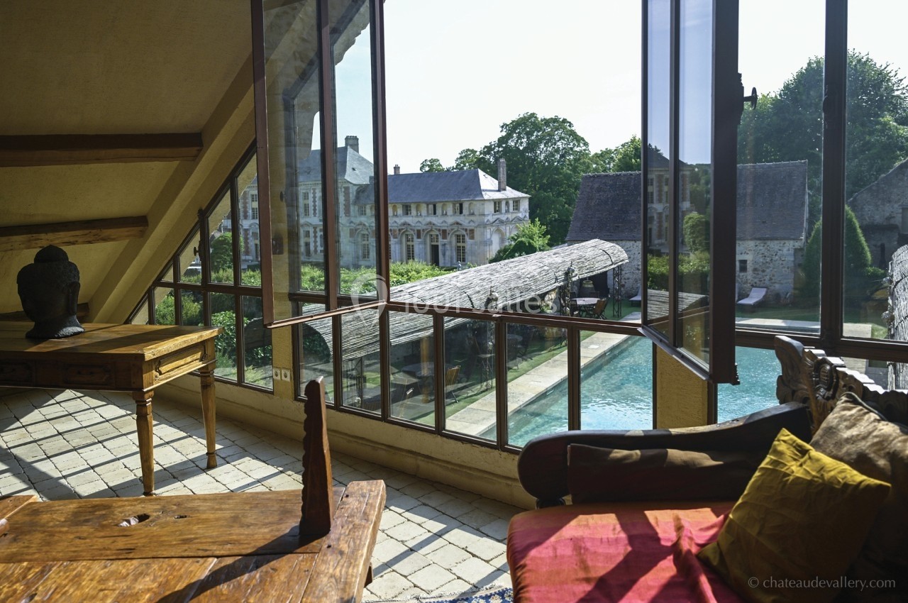 Vue depuis une pièce lumineuse avec mobilier en bois, donnant sur une cour, une piscine et des bâtiments anciens.