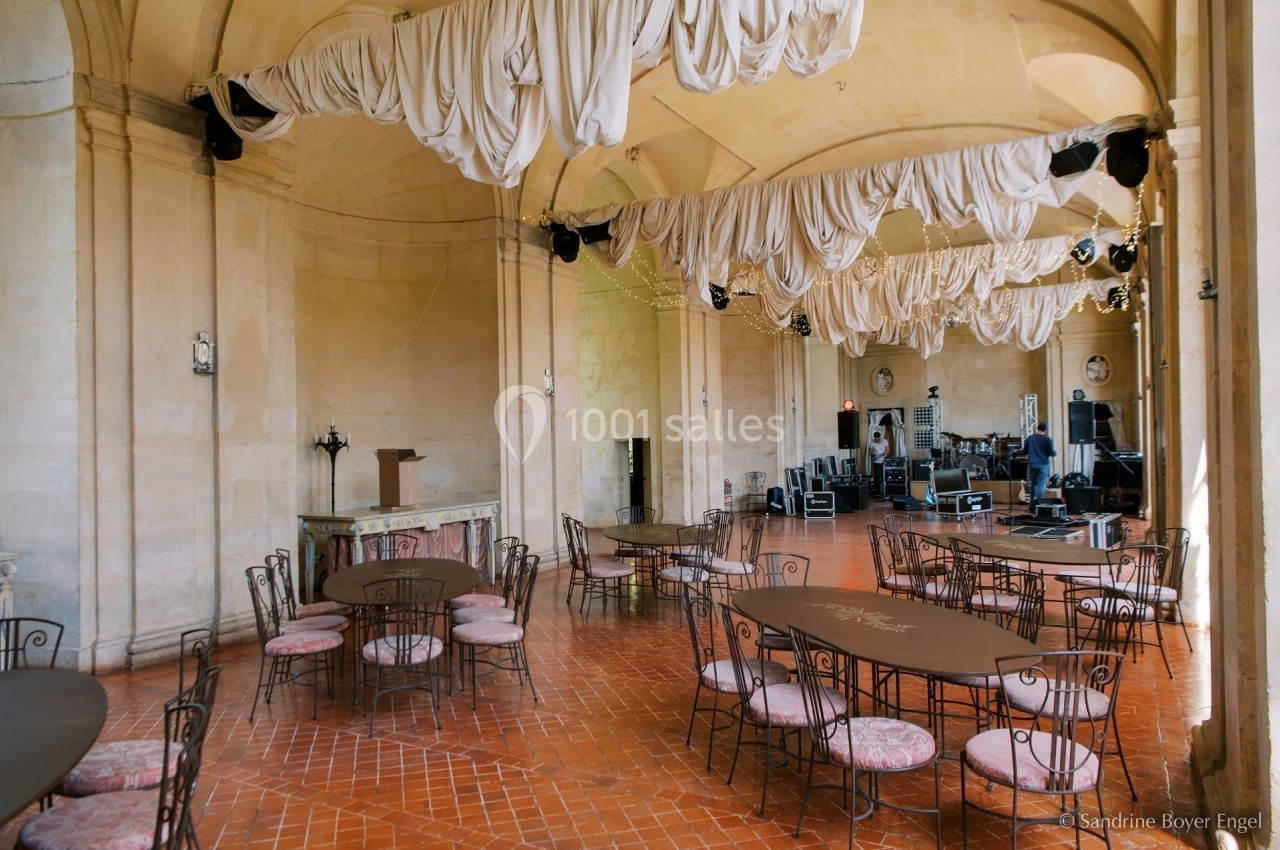 Salle voûtée avec tables et chaises en fer forgé, rideaux suspendus au plafond et scène équipée en arrière-plan.