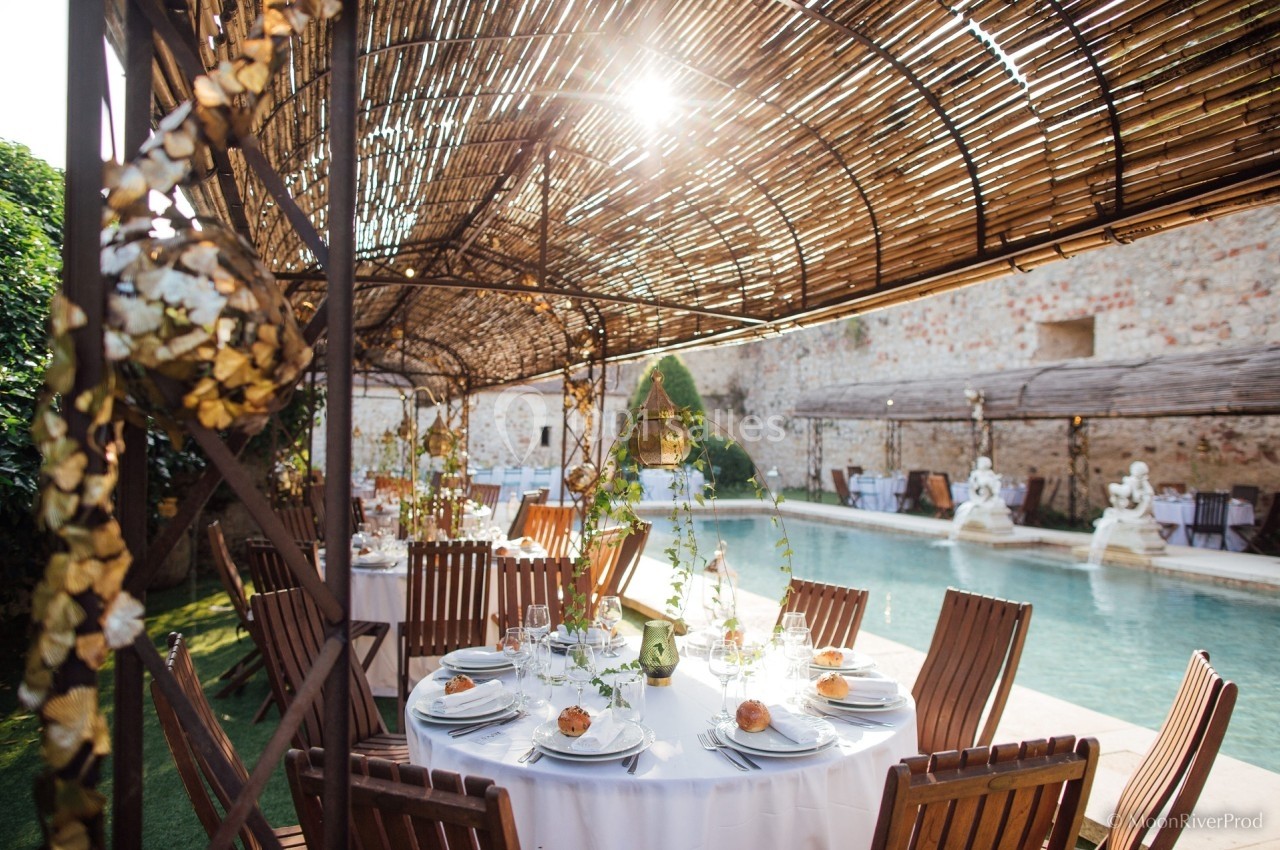 Tables dressées avec élégance sous une pergola en bois, près d'une piscine dans une cour ensoleillée.