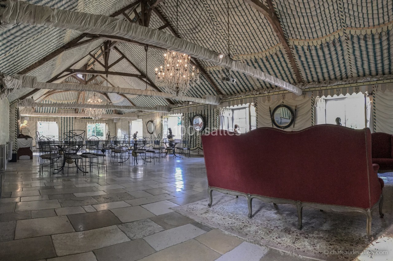 Salle spacieuse avec plafond en poutres apparentes, lustres, mobilier en fer forgé et canapé rouge sur un tapis.