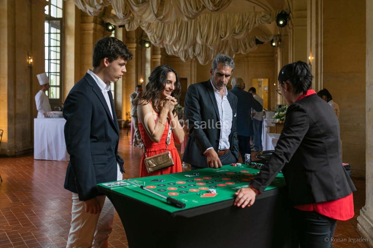 Des personnes jouent à une table de casino dans une salle élégante avec un sol en carreaux rouges et des rideaux suspendus.