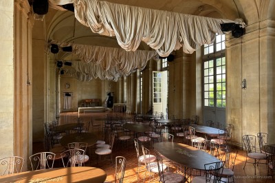 Salle lumineuse avec grandes baies vitrées, tables rondes en métal et lustres suspendus sous un plafond décoré.