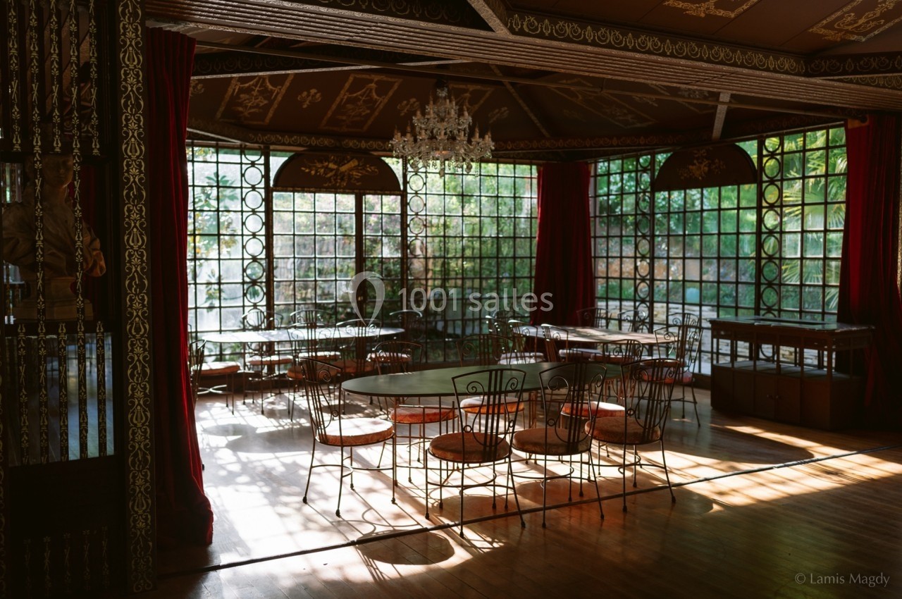 Salle lumineuse avec grandes baies vitrées, rideaux rouges, tables rondes et chaises en métal, décorée d'un lustre.