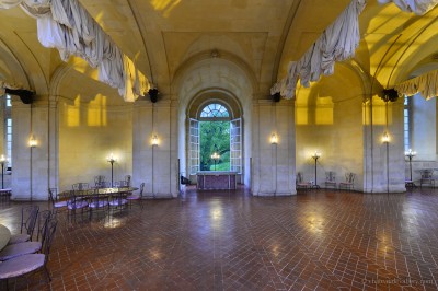 Salle lumineuse avec grandes baies vitrées, tables rondes en métal et lustres suspendus sous un plafond décoré.