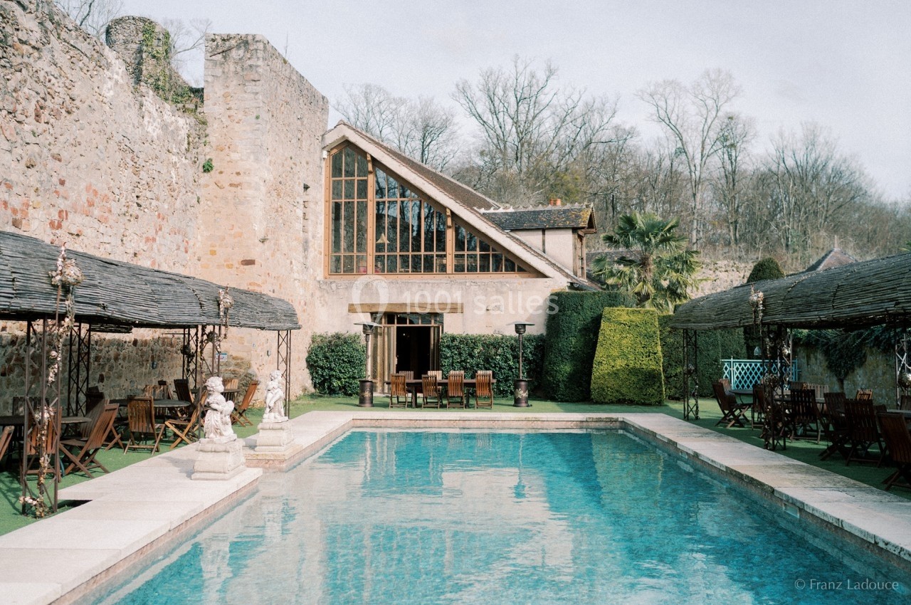 Piscine extérieure bordée de chaises et tables en bois, entourée de murs en pierre et végétation.