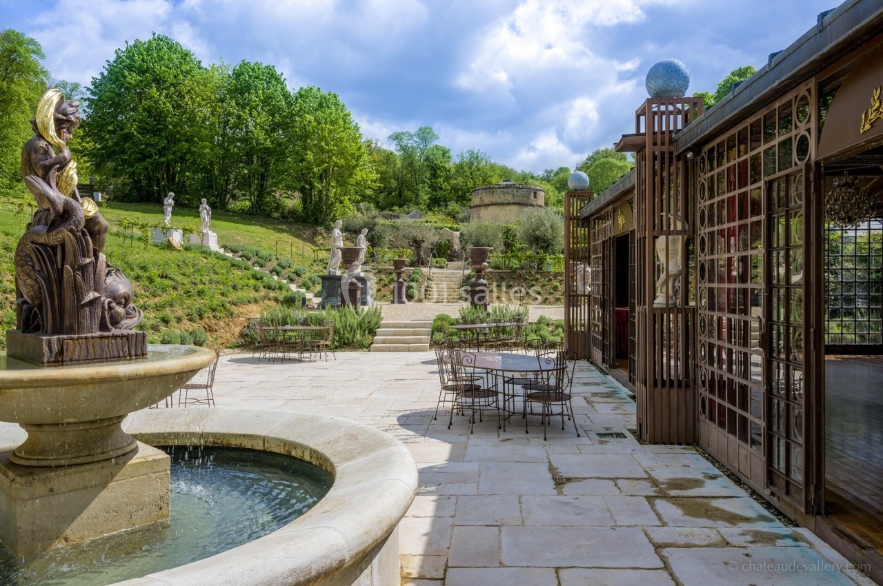 Terrasse pavée avec fontaine, tables en fer forgé, statues et jardin verdoyant sous un ciel partiellement nuageux.