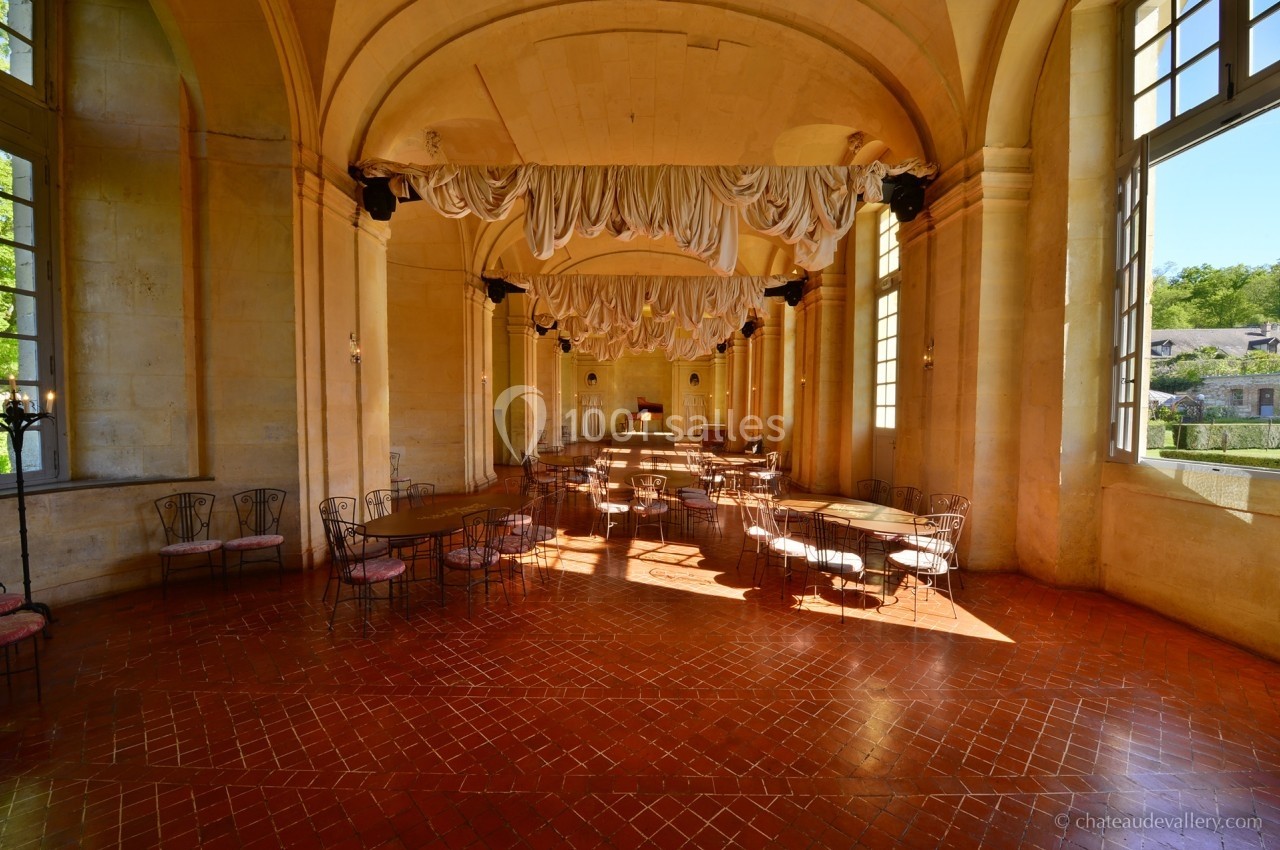 Salle lumineuse avec de grandes fenêtres, sol en terre cuite, chaises en fer forgé et drapés suspendus au plafond.