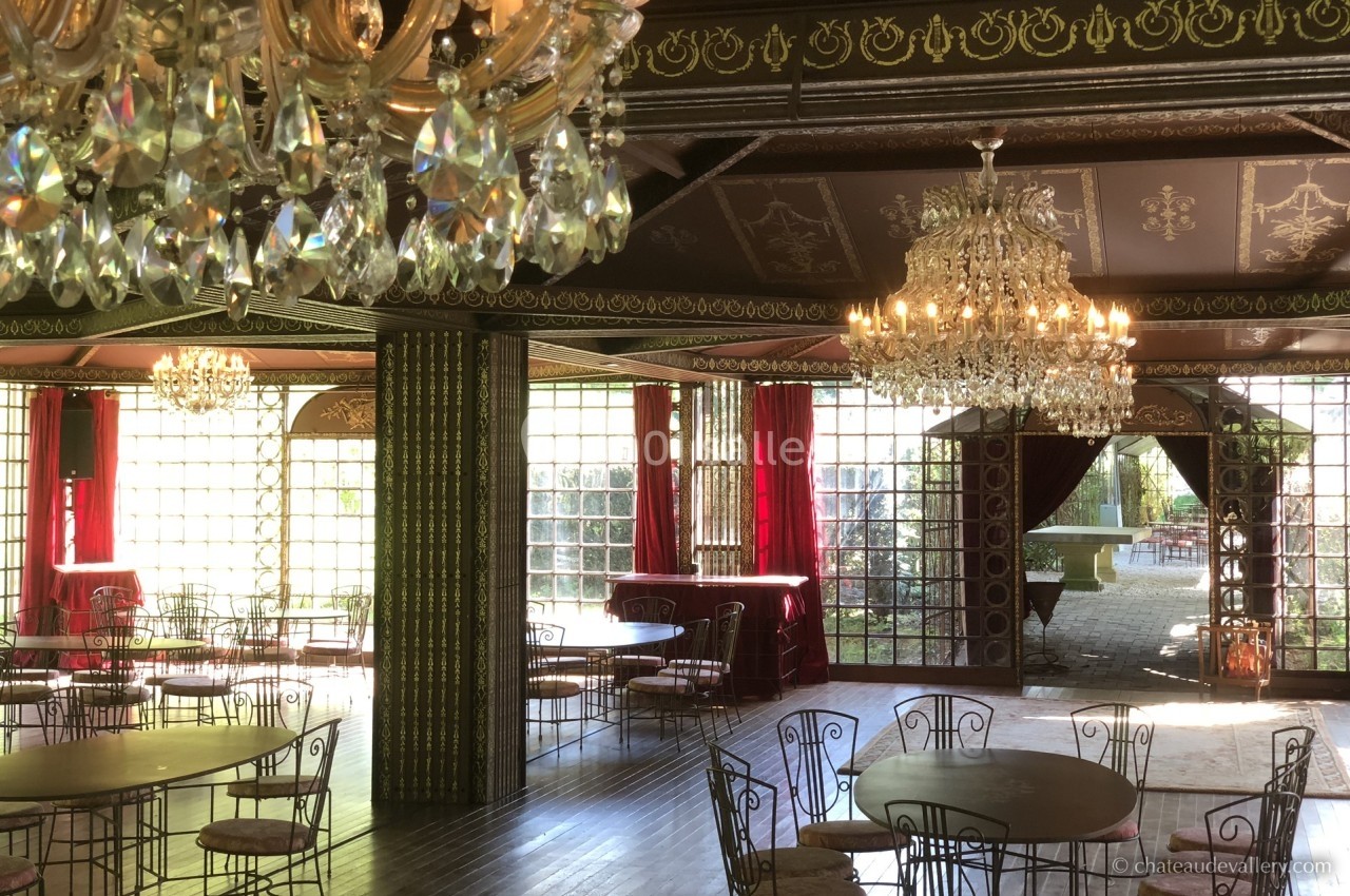 Salle ornée de lustres en cristal, avec des tables et chaises en métal, entourée de grandes baies vitrées.