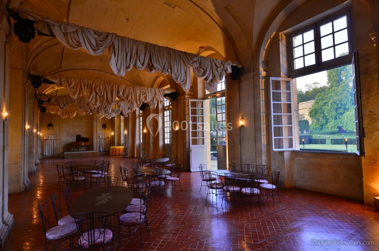 Salle spacieuse avec tables en fer forgé, rideaux suspendus, grandes fenêtres ouvertes sur un jardin verdoyant.