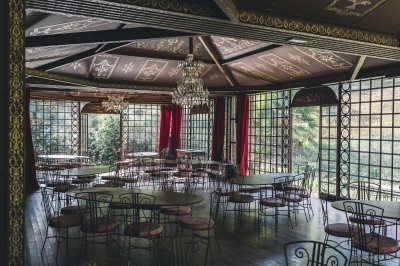 Salle lumineuse avec grandes baies vitrées, tables rondes en métal et lustres suspendus sous un plafond décoré.