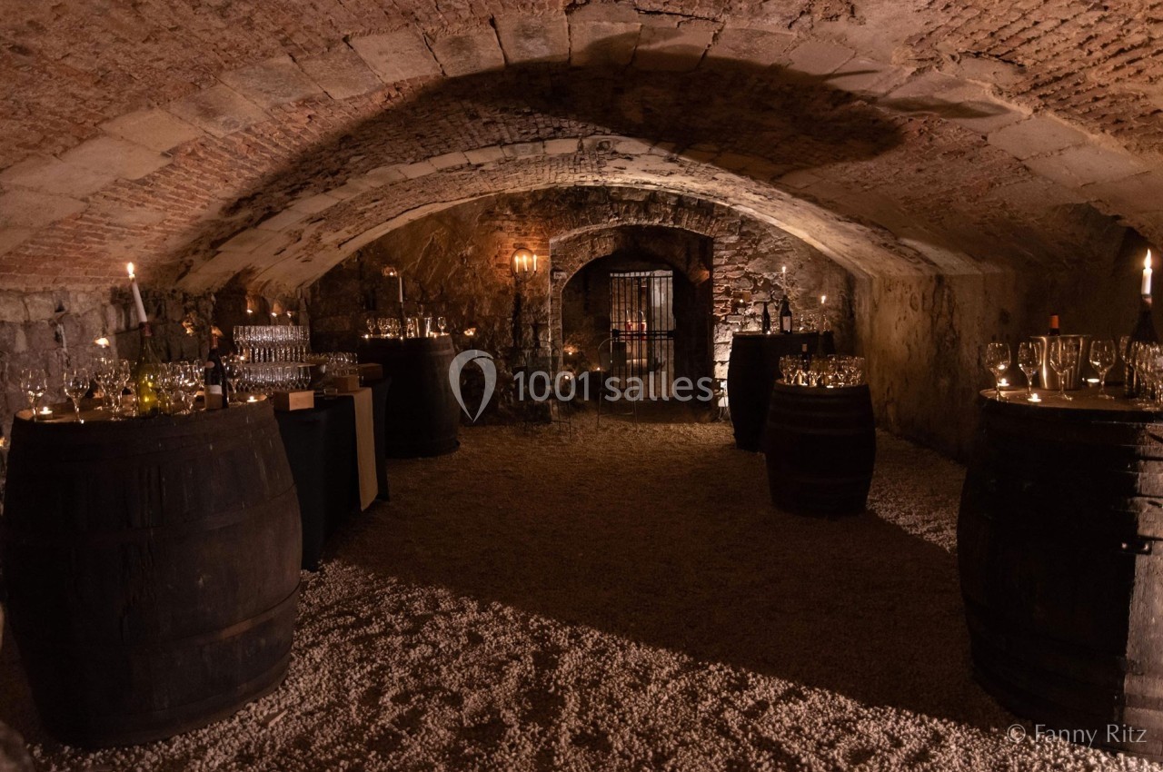 Cave voûtée en pierre éclairée par des bougies, avec des tonneaux et des tables garnies de verres et bouteilles.